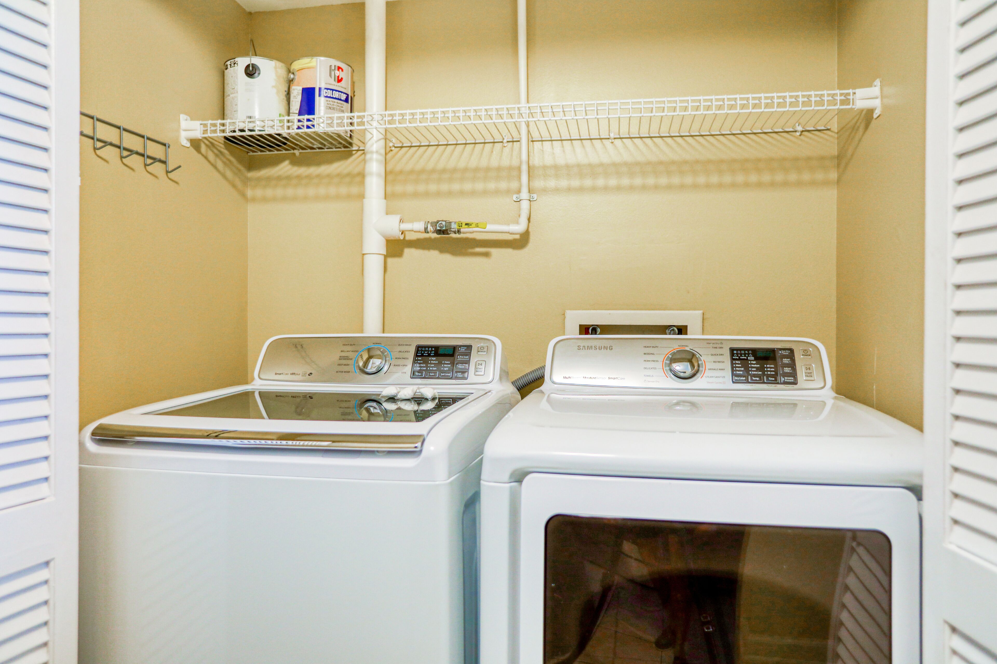 A laundry room with two white washers and dryers, a shelf with cleaning supplies, and a pipe running along the wall.