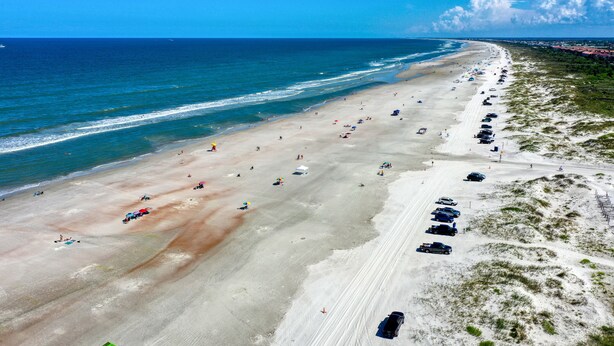 Eine Vogelperspektive eines Strandes mit einer weiten Sandfläche, einigen Sonnenschirmen und geparkten Autos am Ufer.