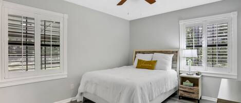 A bedroom with a bed, a nightstand, a ceiling fan, and two windows with blinds.