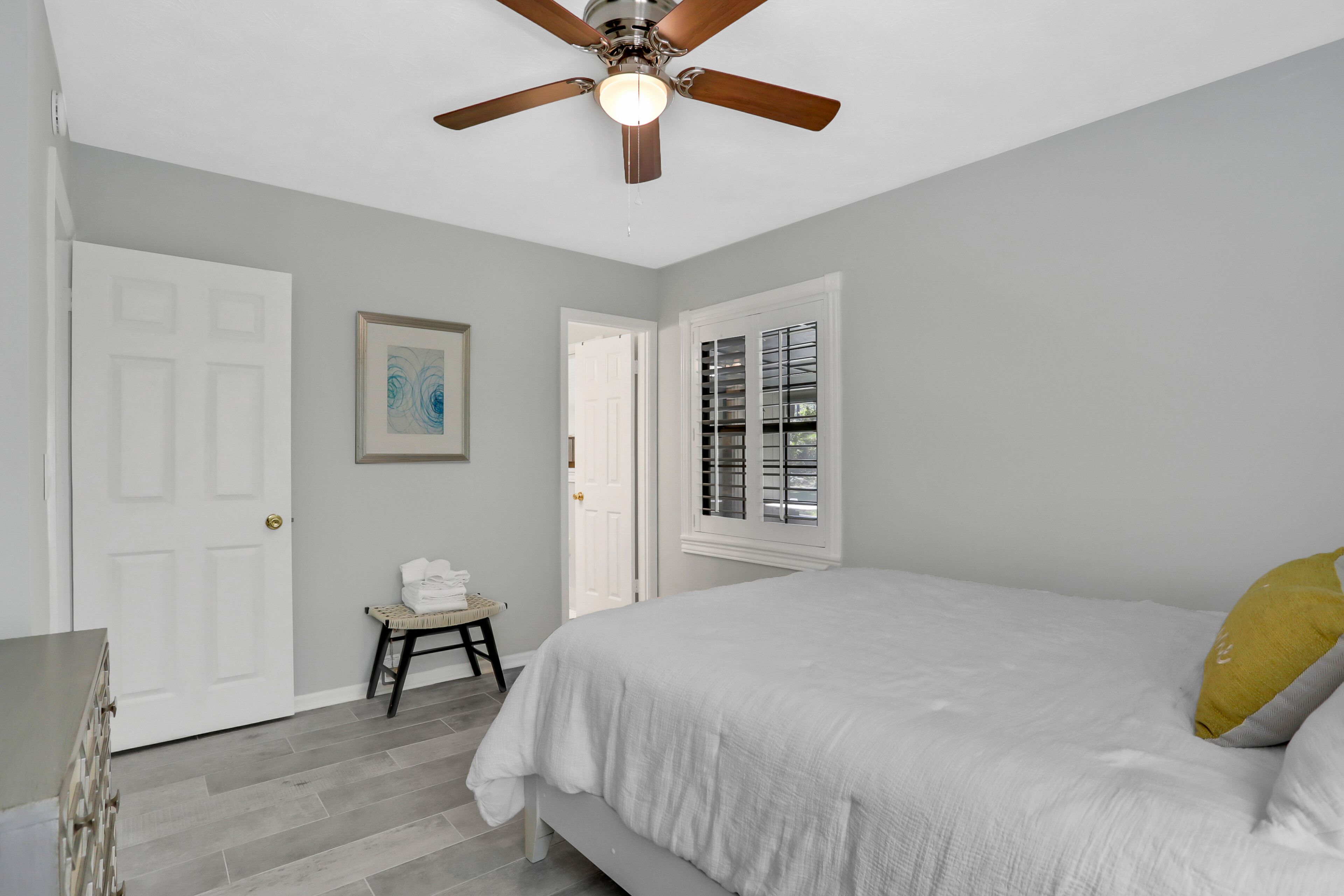 A bedroom with a bed, a ceiling fan, a door, a window with blinds, and a small table with a towel.