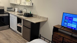 Eine kompakte Küche mit Mikrowelle, Toaster, Spüle und kleinem Kühlschrank. Ein Fernseher ist auf einem Holzständer montiert und zeigt verschiedene Streaming-Dienste.