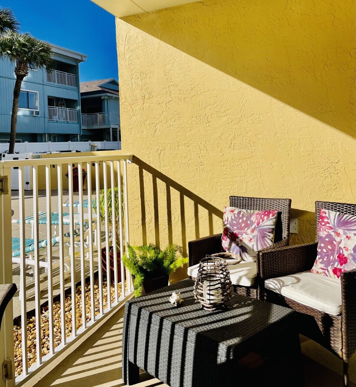 A balcony with wicker furniture, a potted plant, and a view of a pool and buildings.