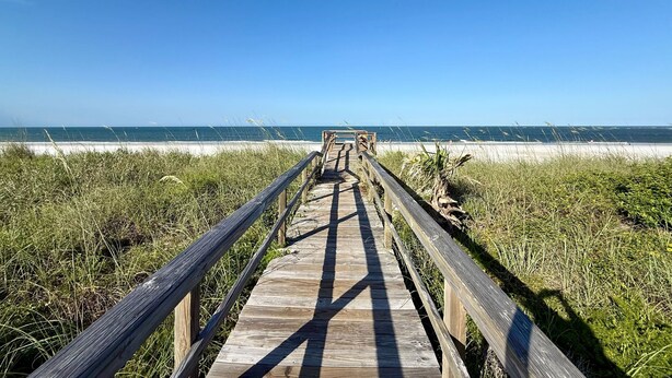 Ein hölzerner Steg führt zu einem Strand unter blauem Himmel.