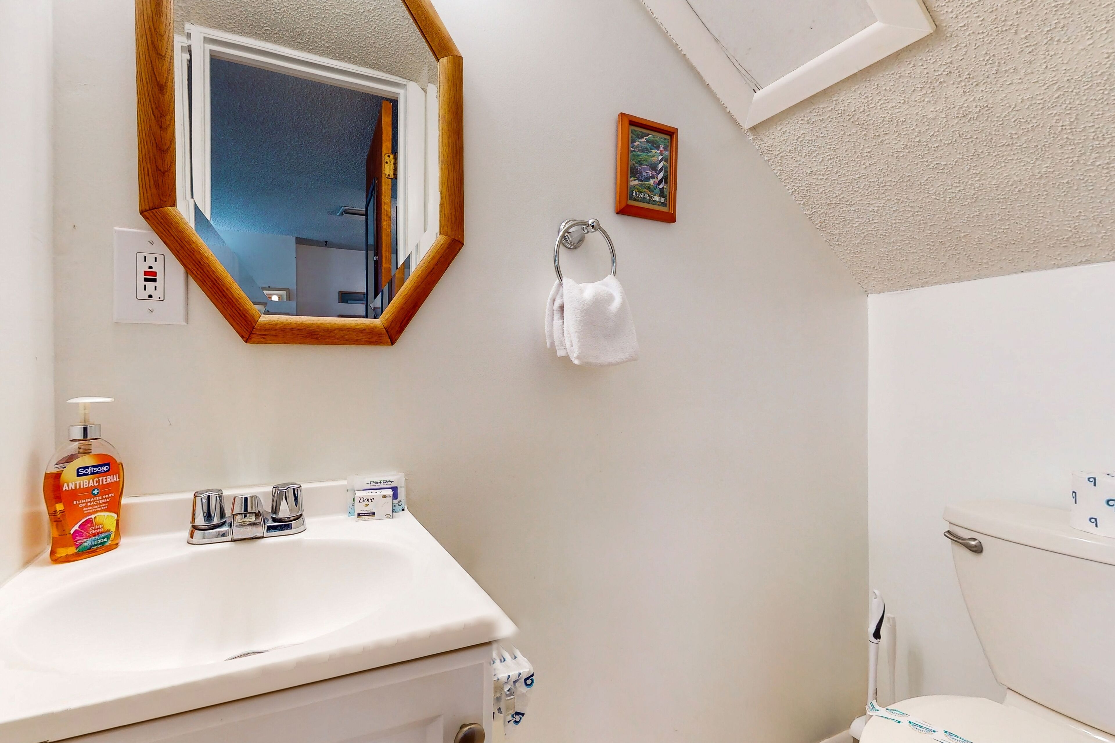 Une salle de bain avec un lavabo blanc, un miroir avec un cadre en bois, un petit tableau encadré accroché au mur et des toilettes avec un porte-rouleau de papier toilette.