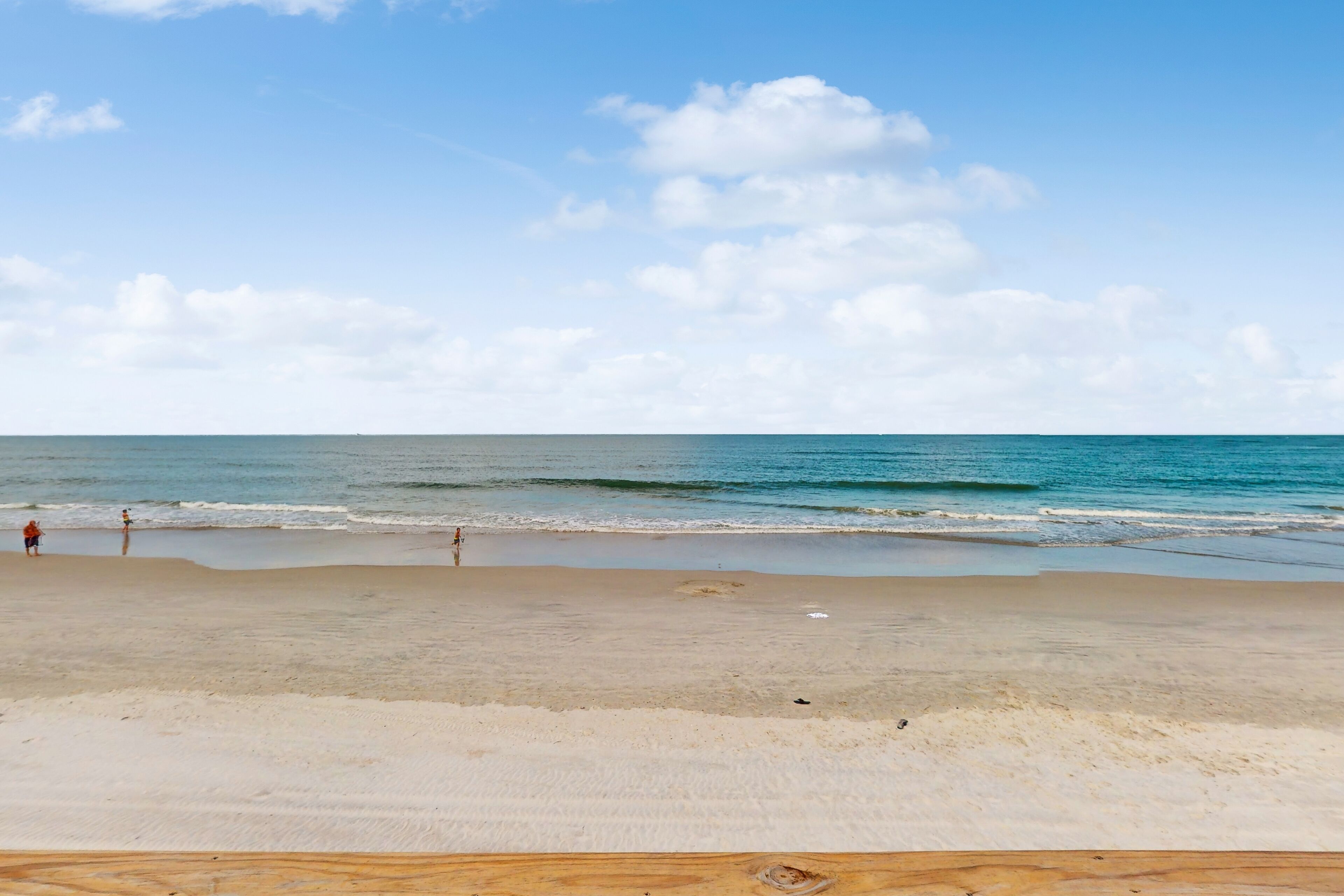 A beach with a wooden deck in the foreground, a sandy shore, and a clear sky with clouds.