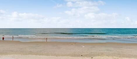 A beach with a wooden deck in the foreground, a sandy shore, and a clear sky with clouds.