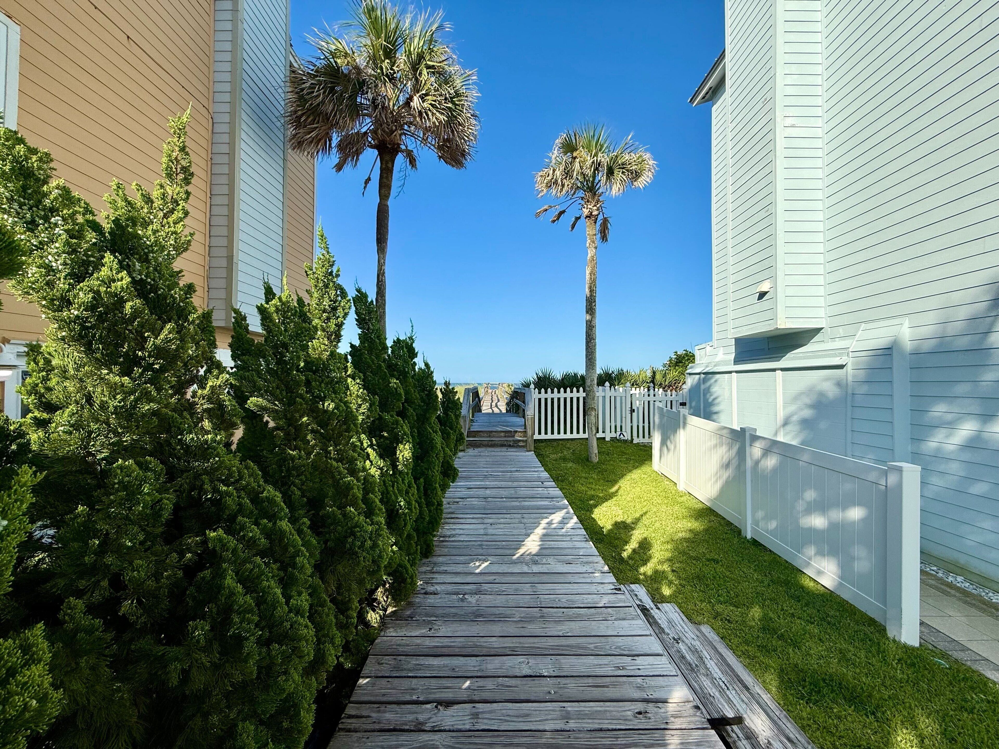 A wooden pathway flanked by green shrubs and palm trees, leading to a white picket fence.