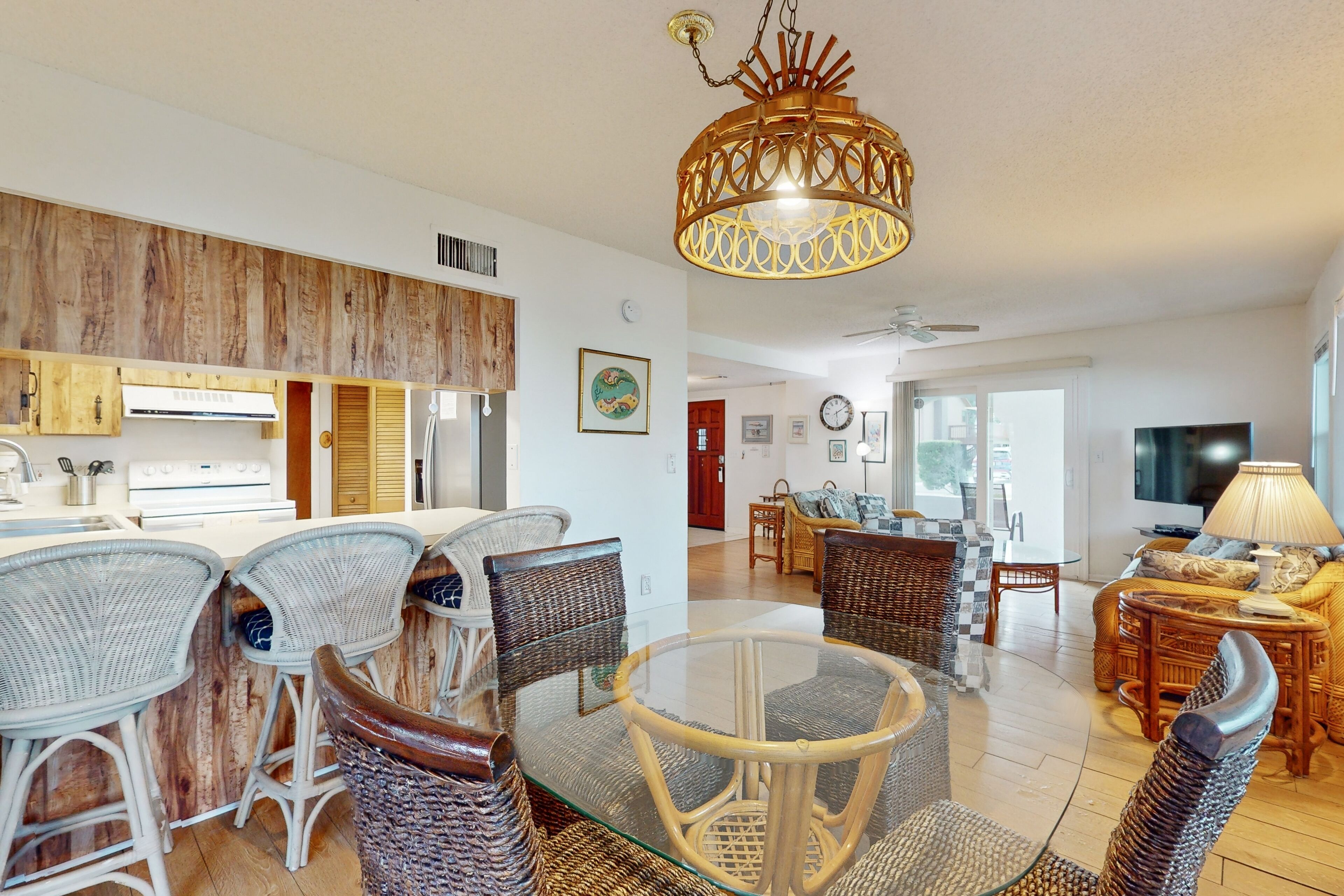 A modern kitchen with white appliances, a dining area with wicker furniture, and a living room with a TV and sofa.