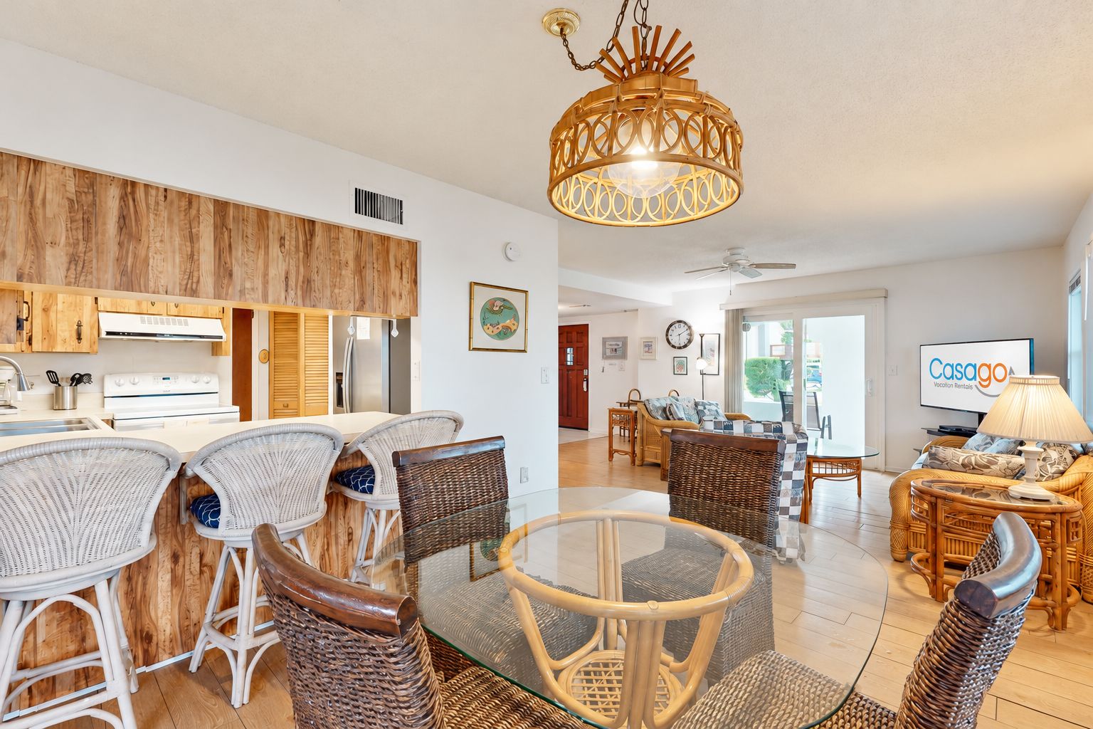 A modern kitchen with white appliances, a dining area with wicker chairs, a glass-top table, and a living room with a sofa and a television.