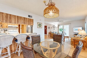 A modern kitchen with white appliances, a dining area with wicker chairs, a glass-top table, and a living room with a sofa and a television. - Spacious Seaside Cottage w/ Sunroom 1-minute Walk to Vilano Beach (St. Augustine)