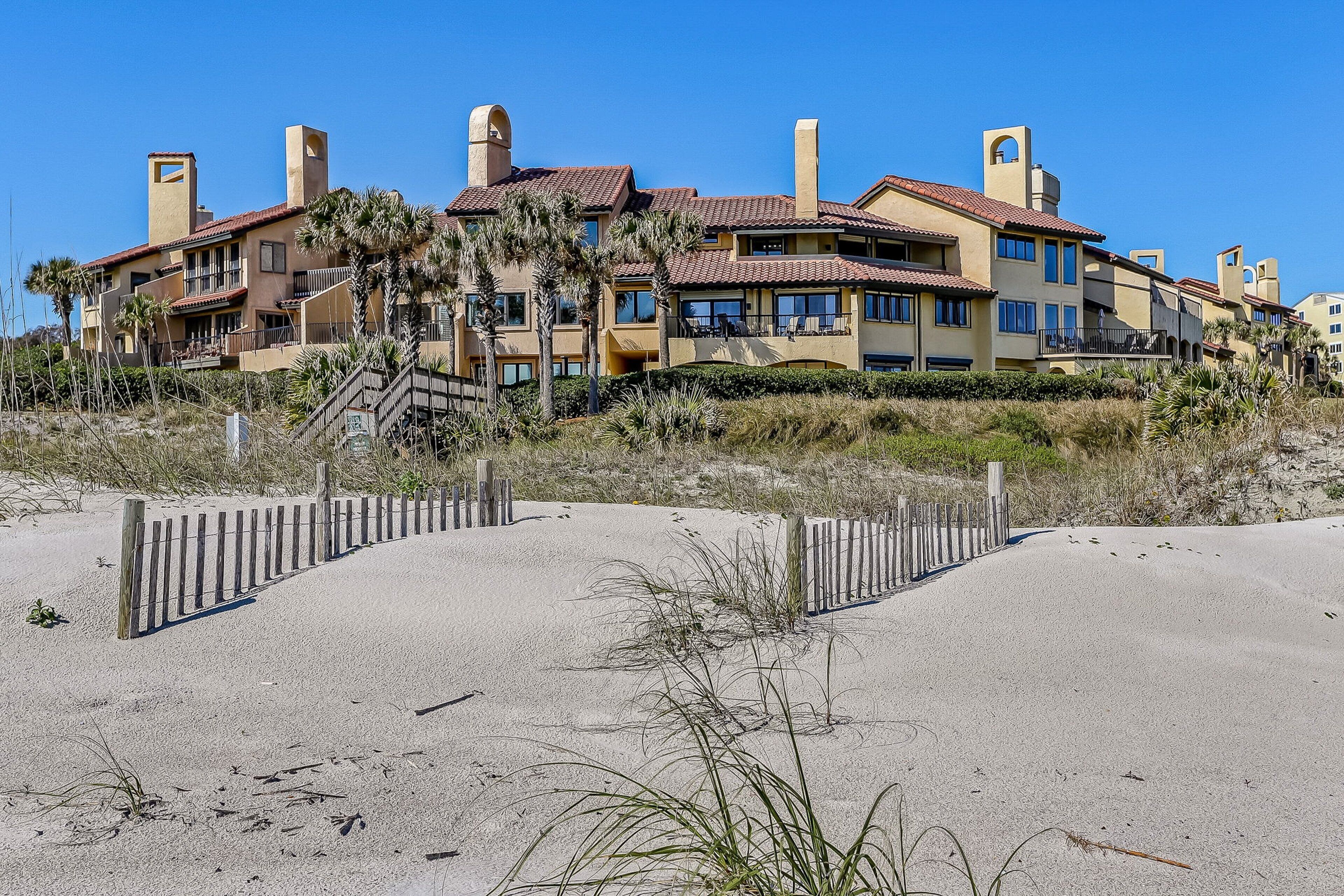 A beachfront hotel with multiple balconies, palm trees, and a sandy beach in the foreground.
