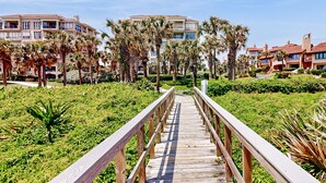 A wooden boardwalk leading to a building complex with multiple balconies and palm trees. - Snowbird Friendly, Oceanfront Community With Optional Golf Cart! (Fernandina Beach)