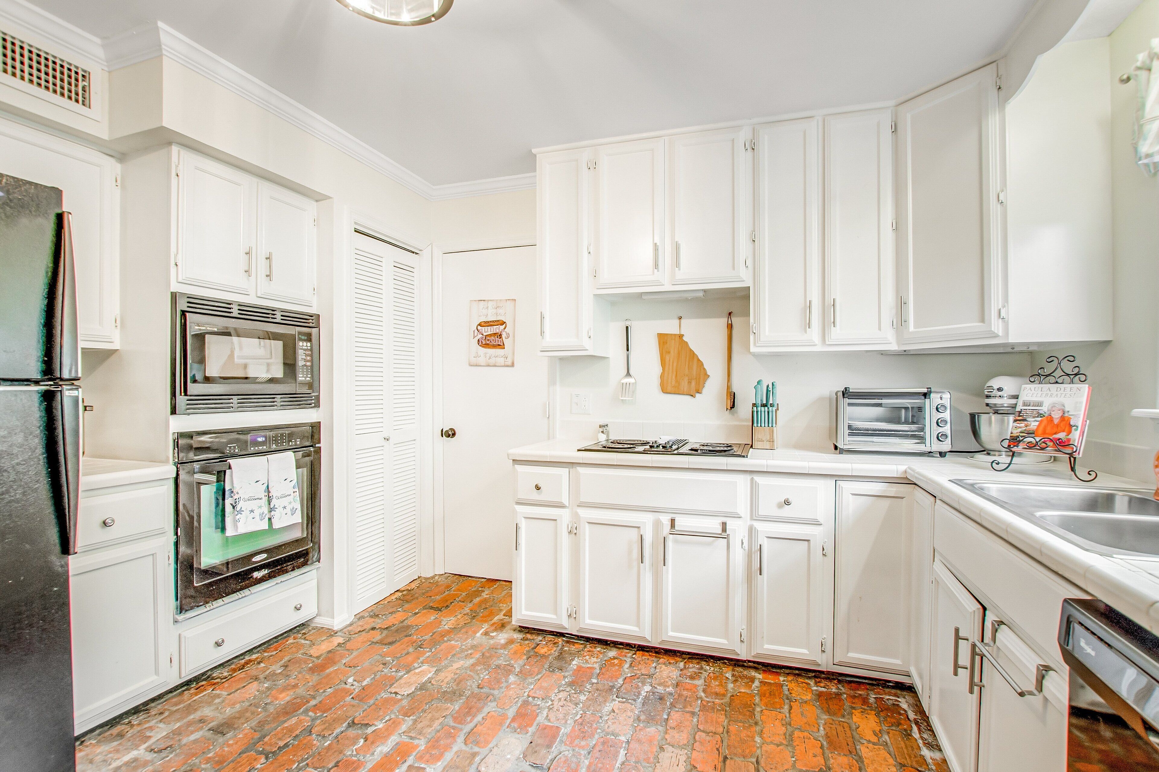 A kitchen with white cabinets, a brick floor, a refrigerator, an oven, a toaster, and a sink.
