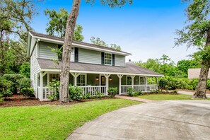 A two-story house with a porch, surrounded by trees and a well-maintained lawn. - Relaxing, Dog-friendly Home With Furnished Patio, Washer/dryer, Wifi, & AC (St. Simons Island)