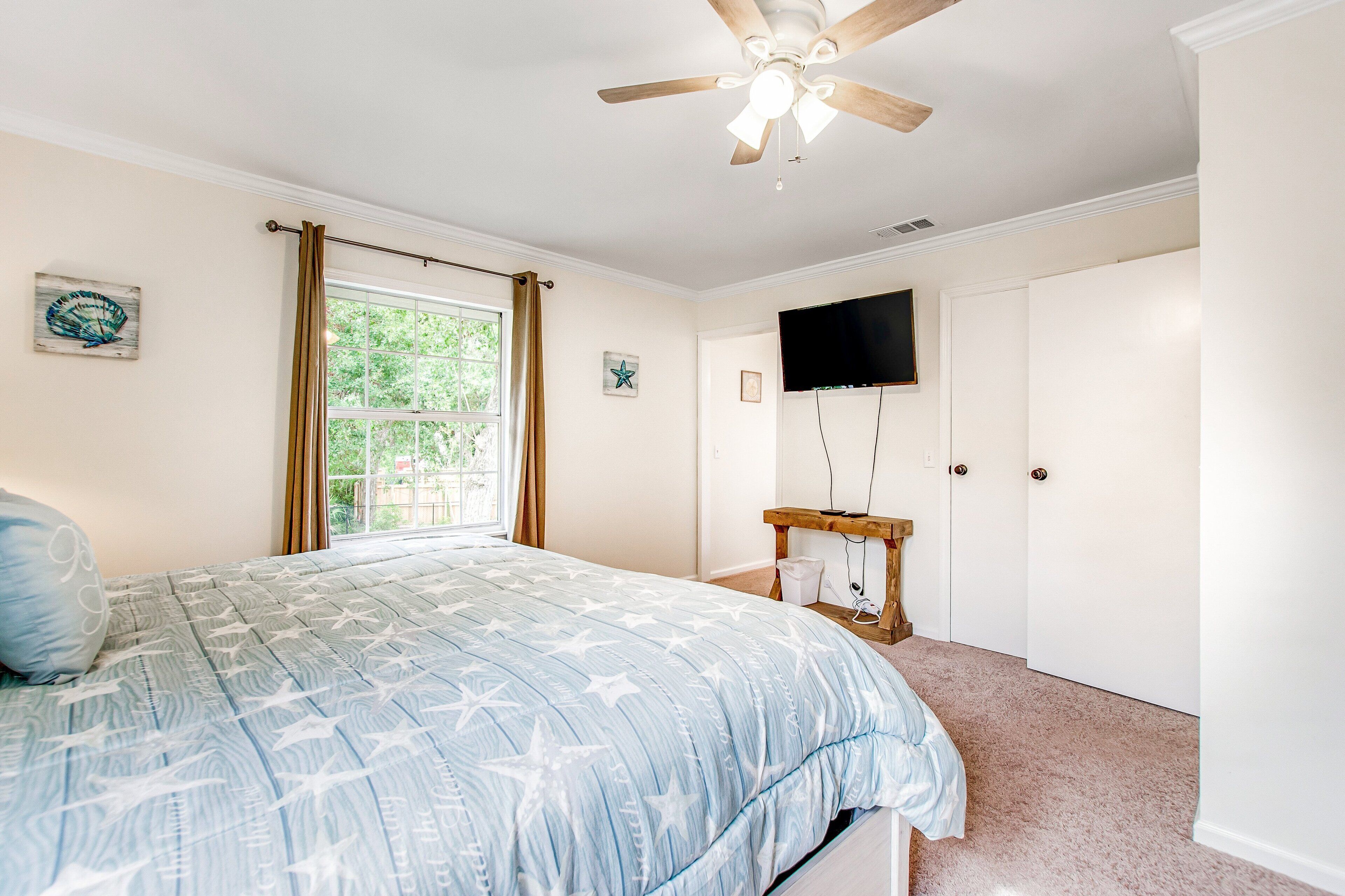 A bedroom with a bed, a ceiling fan, a window with curtains, a television mounted on the wall, and a door.