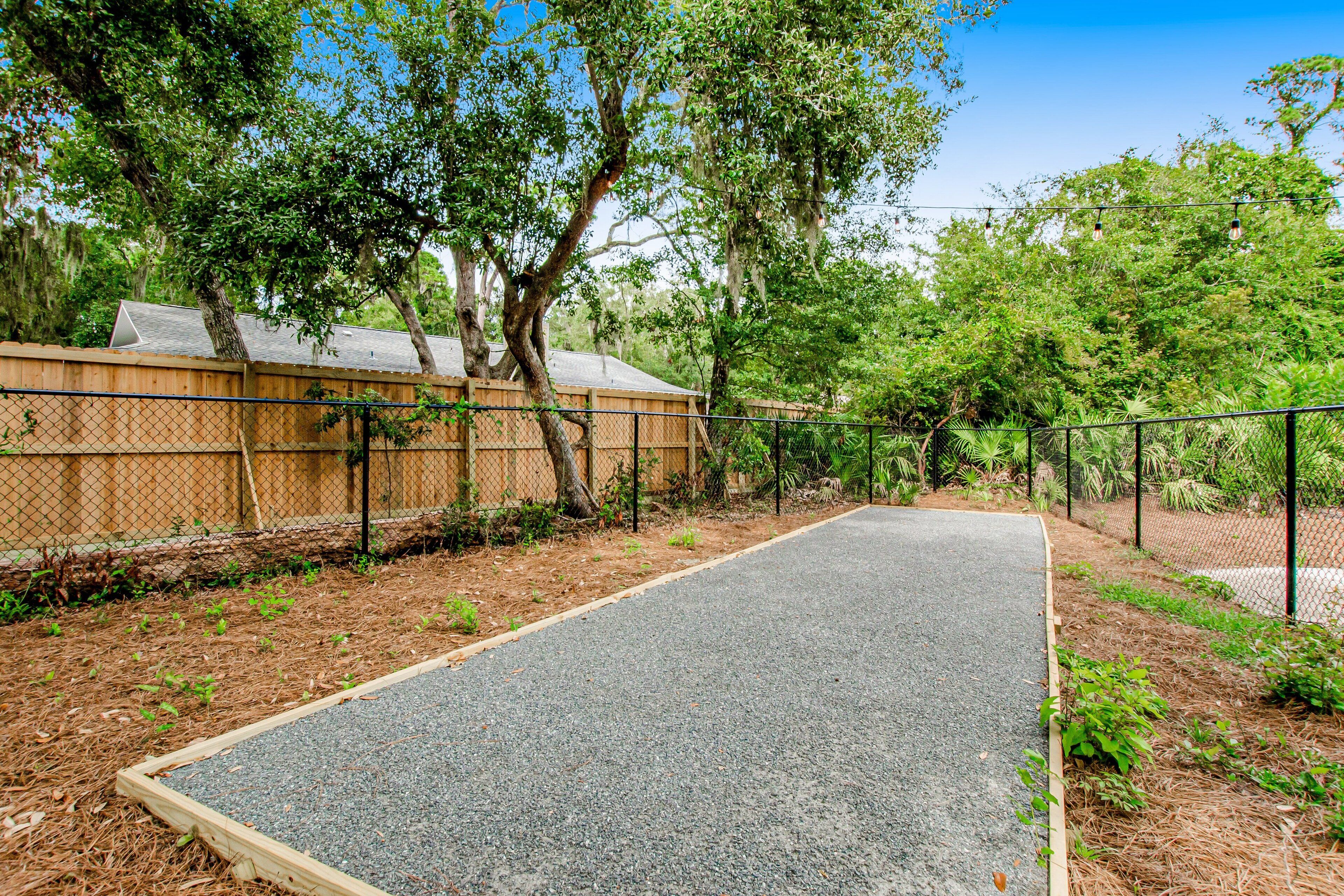 A paved pathway bordered by a wooden fence and trees, leading to a building with a sloped roof.
