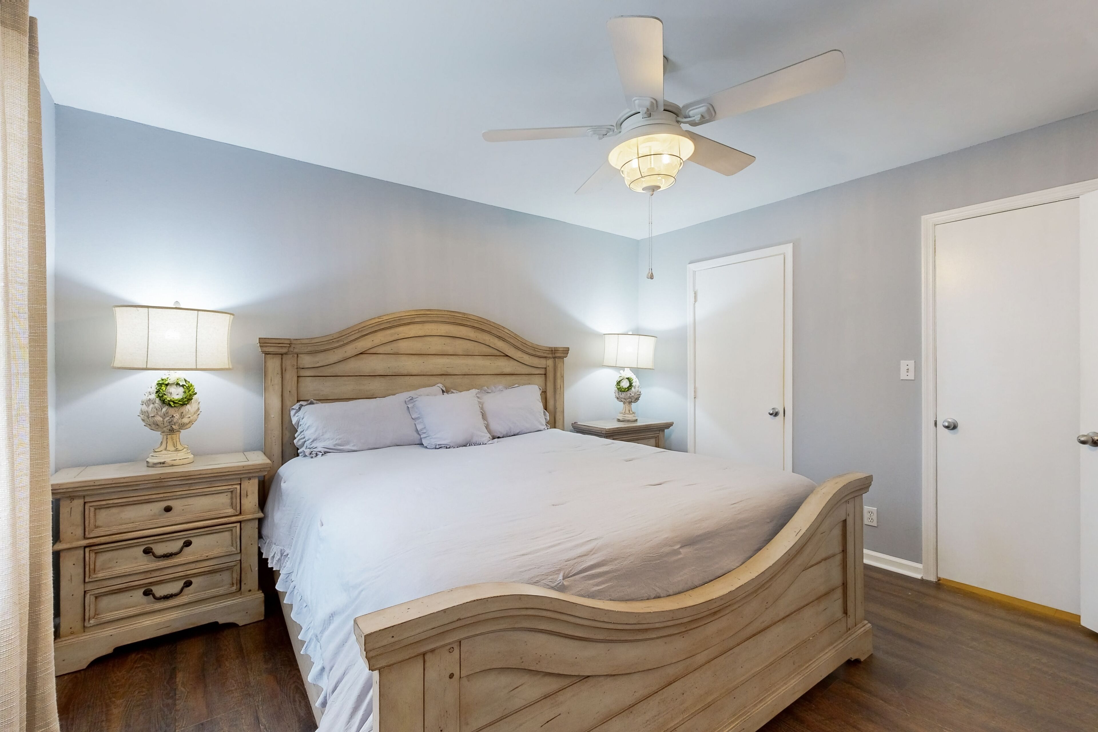 Un dormitorio con una cama de madera, dos mesitas de noche con lámparas, un ventilador de techo y una puerta.