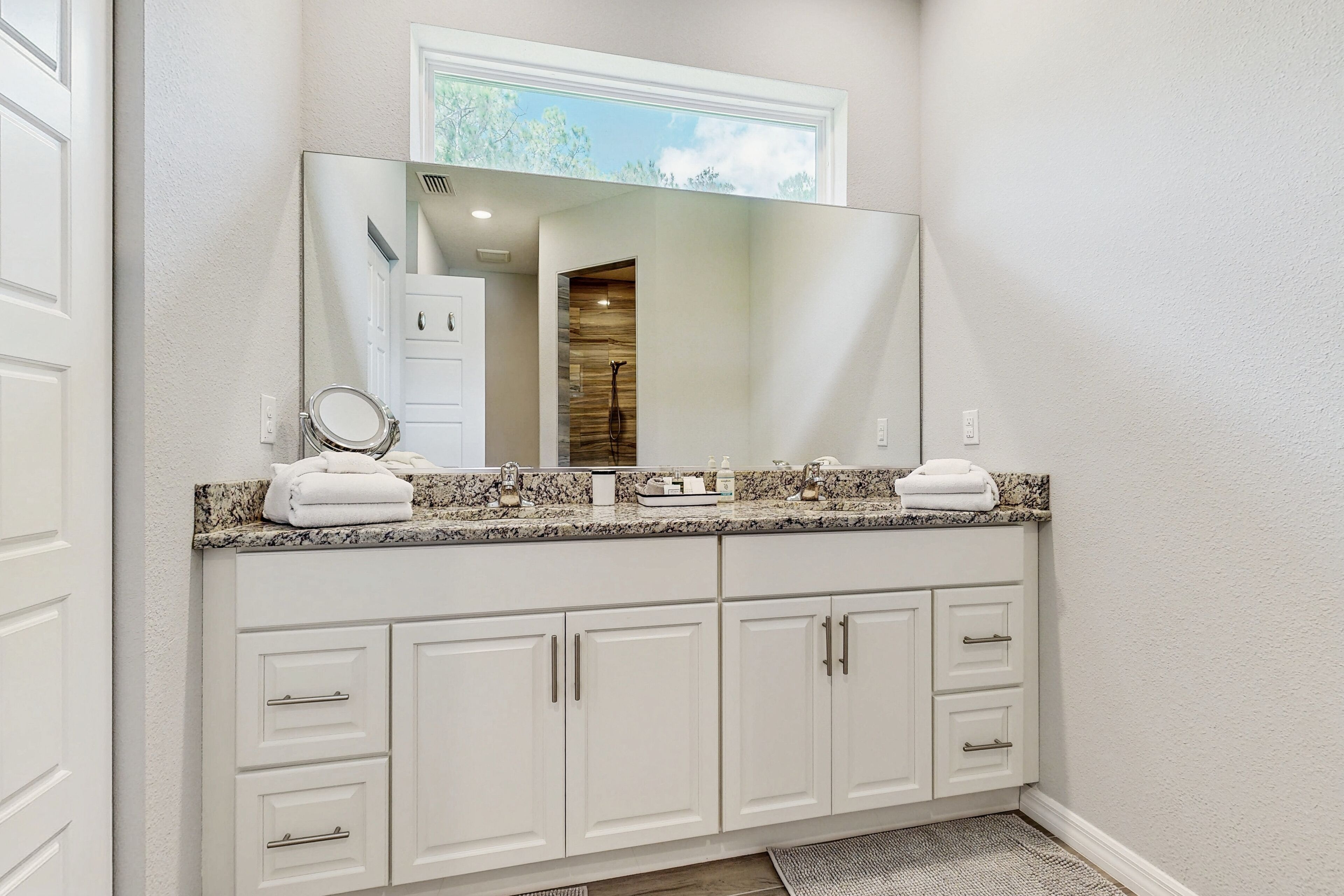 A bathroom with a large mirror, white cabinets, a granite countertop, and a visible shower area.