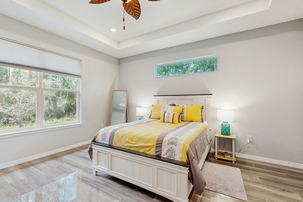 A bedroom with a large window, a bed with a headboard, a bedside table with a lamp, and a wall-mounted artwork. - Spacious House With Private Covered Patio, Work Station, Full Kitchen and Grill (Palm Coast)