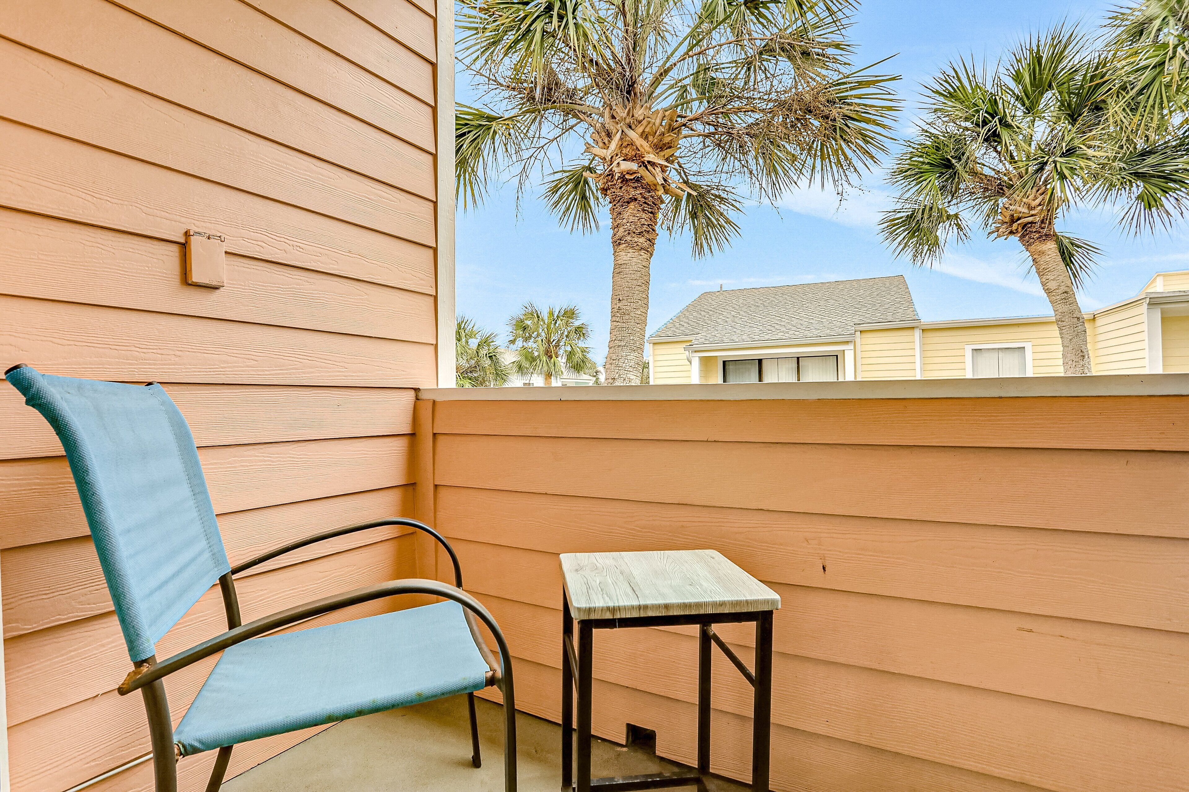 A balcony with a chair and a small table, overlooking palm trees and a house.