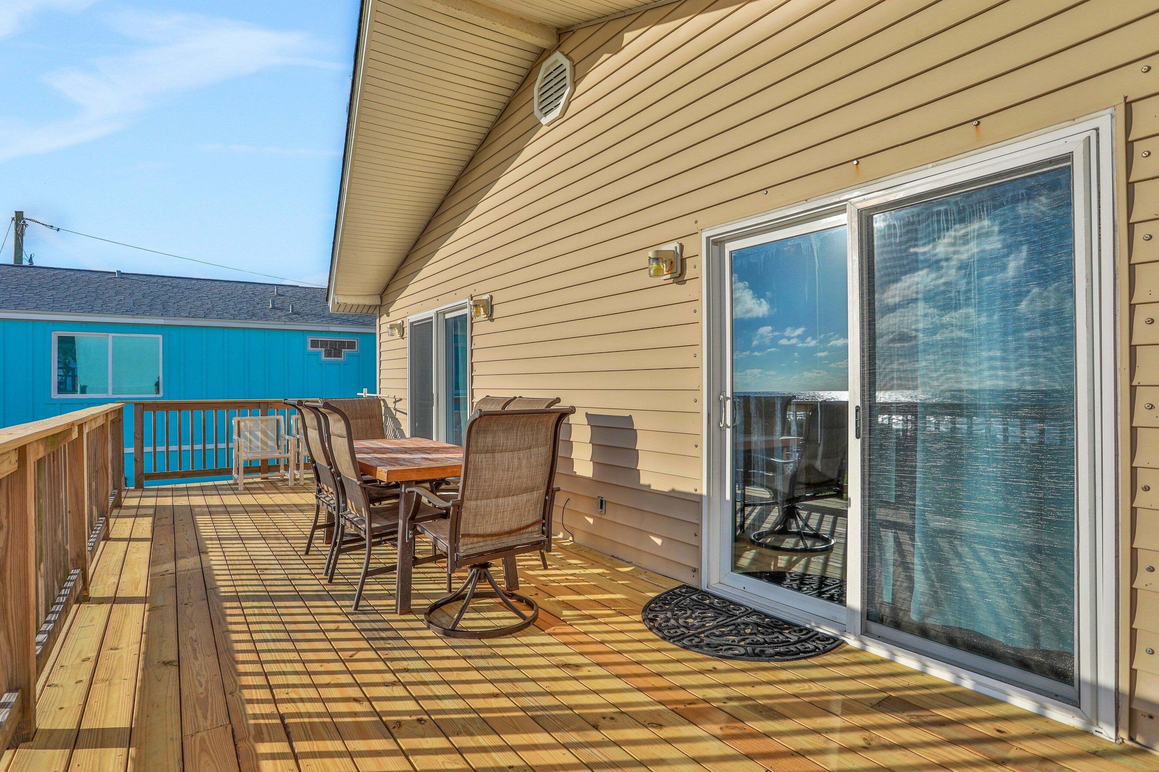 A wooden deck with a table and chairs, a sliding glass door, and a blue building in the background.