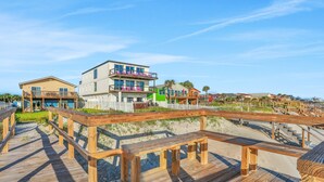 Une promenade en bois mène à une propriété située en bord de mer, qui comprend plusieurs bâtiments, notamment une maison à deux étages dotée de balcons et une autre construction avec un toit végétalisé.