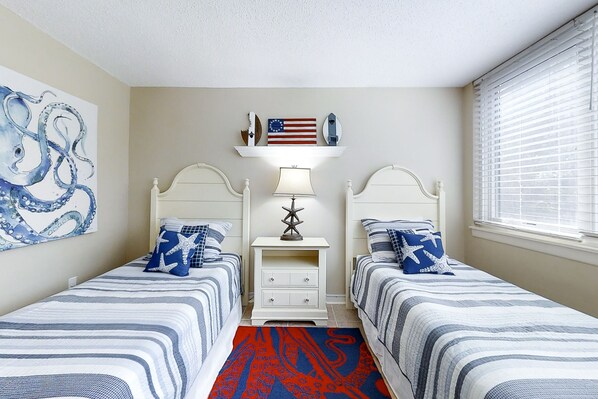 A bedroom with two beds, a nightstand, a lamp, and a wall shelf with decorative items. - Renovated Condo With Balcony, Lush Views, Outdoor Pool (Fernandina Beach)