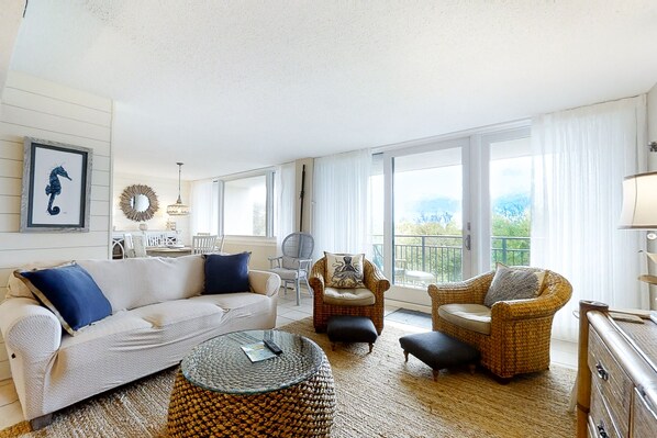 A spacious living room with a white sofa, two armchairs, a round coffee table, and a view of the outdoors through large windows. - Renovated Condo With Balcony, Lush Views, Outdoor Pool (Fernandina Beach)