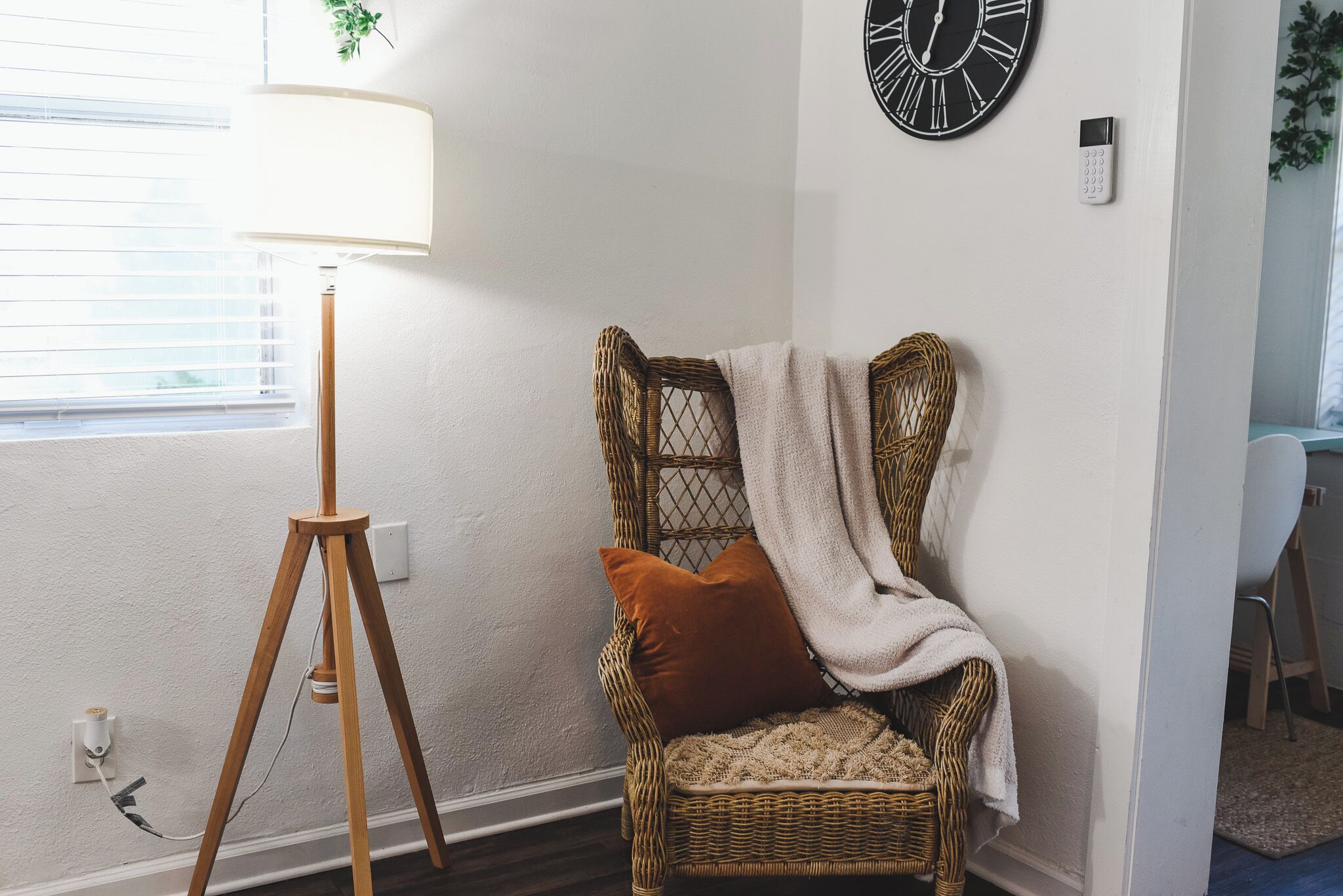 A corner of a room with a wicker chair, a lamp, a clock, and a wall socket.