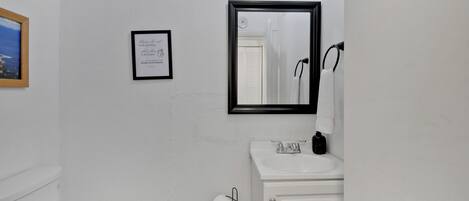 Un baño con inodoro, lavamanos y espejo. Hay un cuadro en la pared y un rollo de papel higiénico.