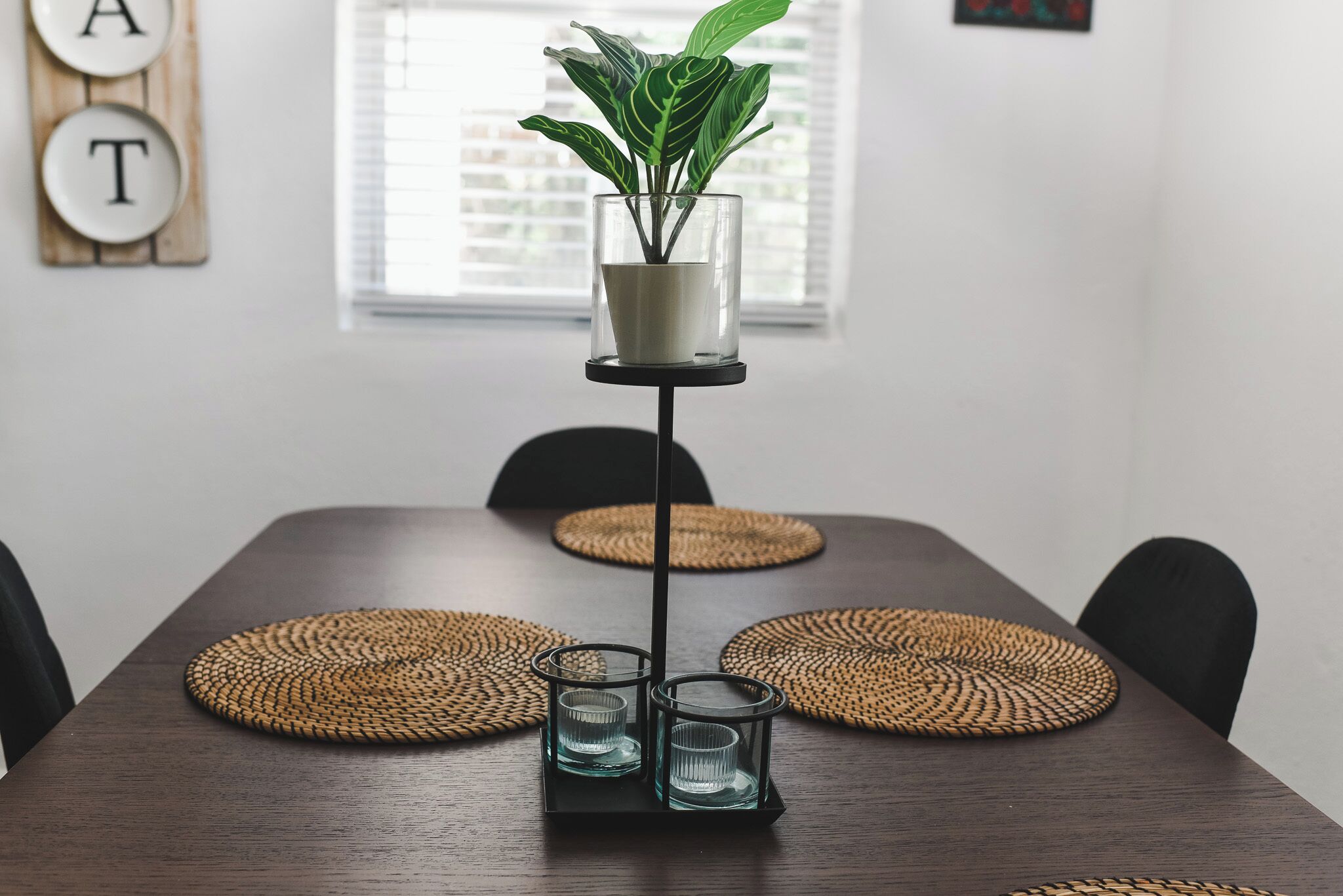 A dining table with a potted plant, three glass candle holders, and woven placemats.