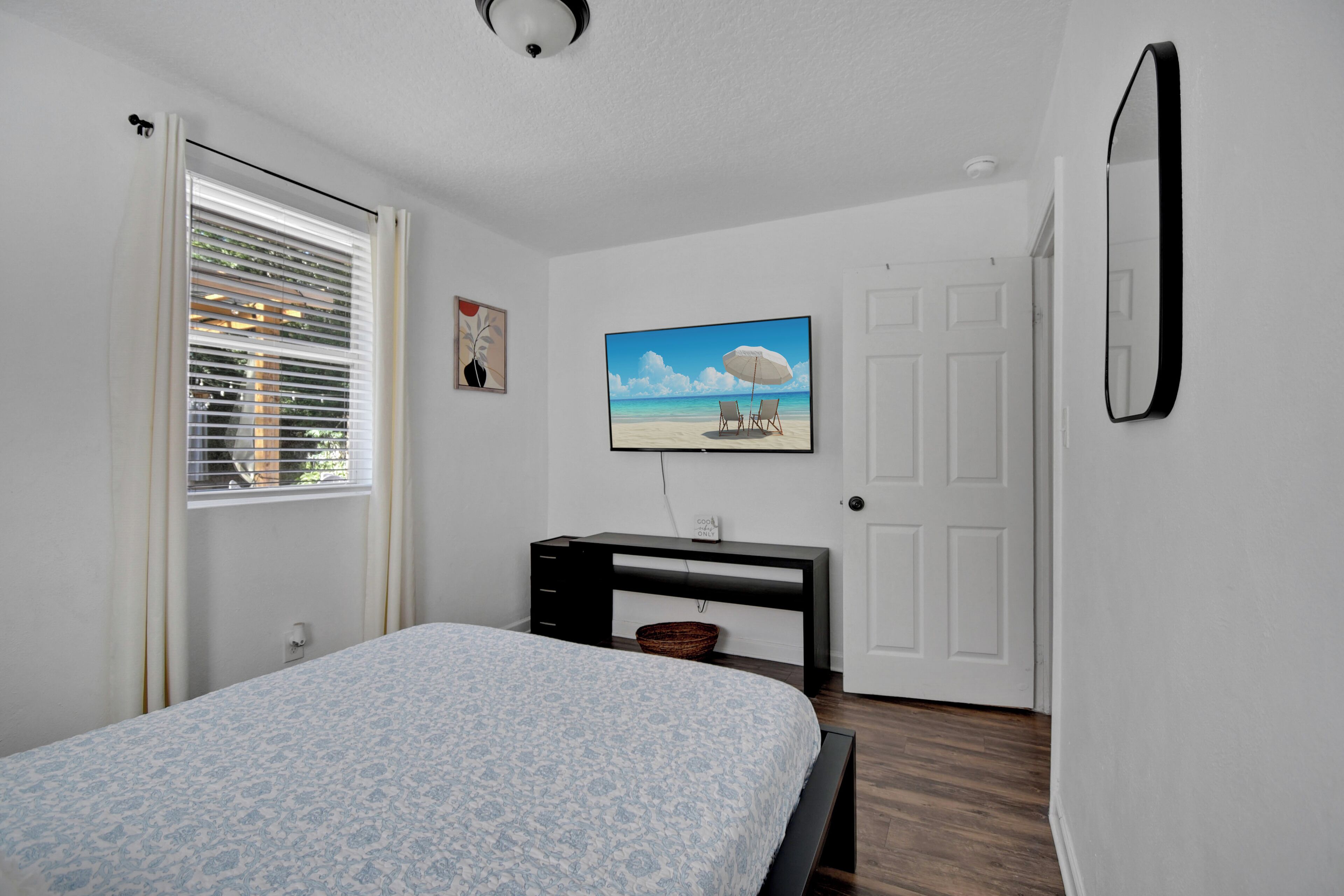 Een slaapkamer met een bed, een bureau, een schilderij van een strand en een deur.