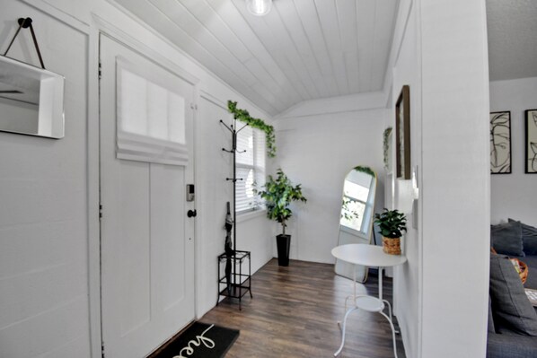 Un couloir avec un plancher en bois, des murs blancs, un porte-manteau, une plante en pot, une petite table et une porte avec un paillasson.