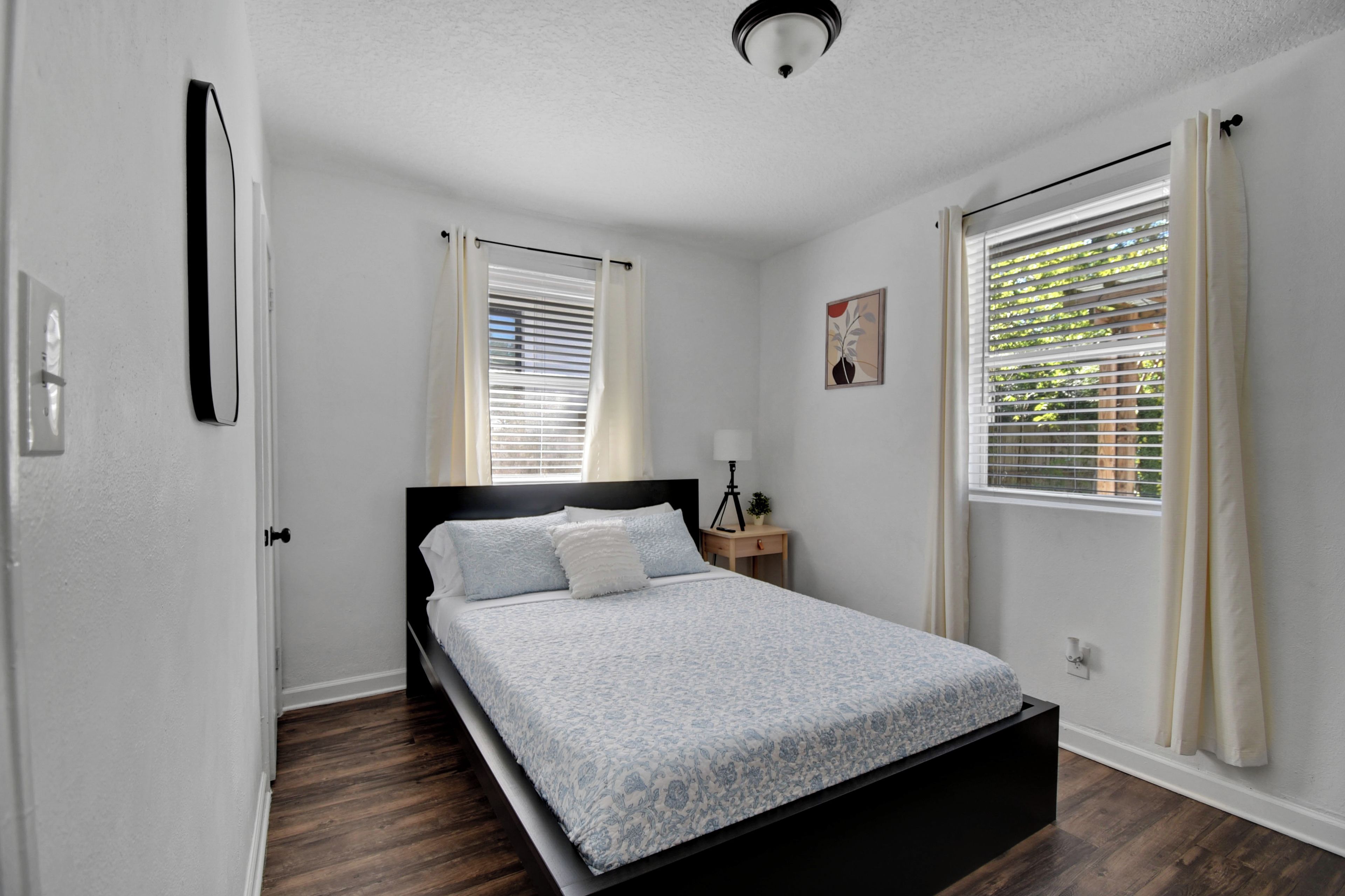 A bedroom with a bed, a nightstand, a lamp, and two windows with curtains.