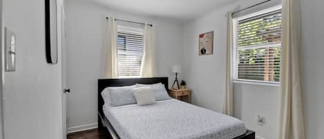 Un dormitorio con cama, mesita de noche, lámpara y dos ventanas con cortinas.
