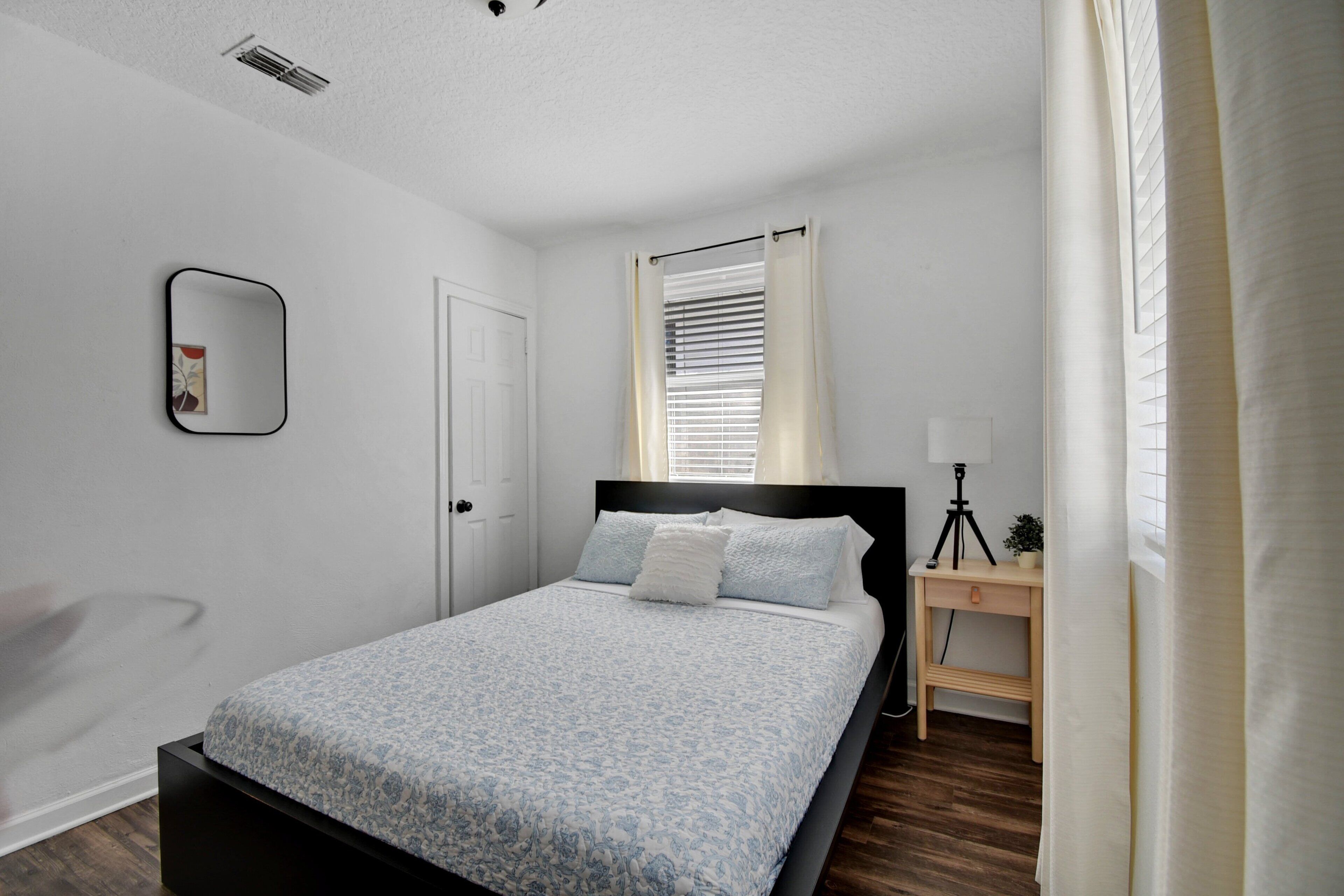 A bedroom with a bed, a bedside table with a lamp, a chair, and a window with blinds.