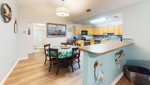 A modern kitchen with wooden cabinets, a dining area with a floral tablecloth, and a light blue accent wall. - Serene Home With Enclosed Yard, Grill, Games, & Central AC - Dog-friendly (St. Augustine)