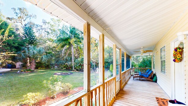 A covered porch with wooden railings, a seating area with blue chairs, and a view of a landscaped garden with various plants and trees. - Serene Home With Enclosed Yard, Grill, Games, & Central AC - Dog-friendly (St. Augustine)