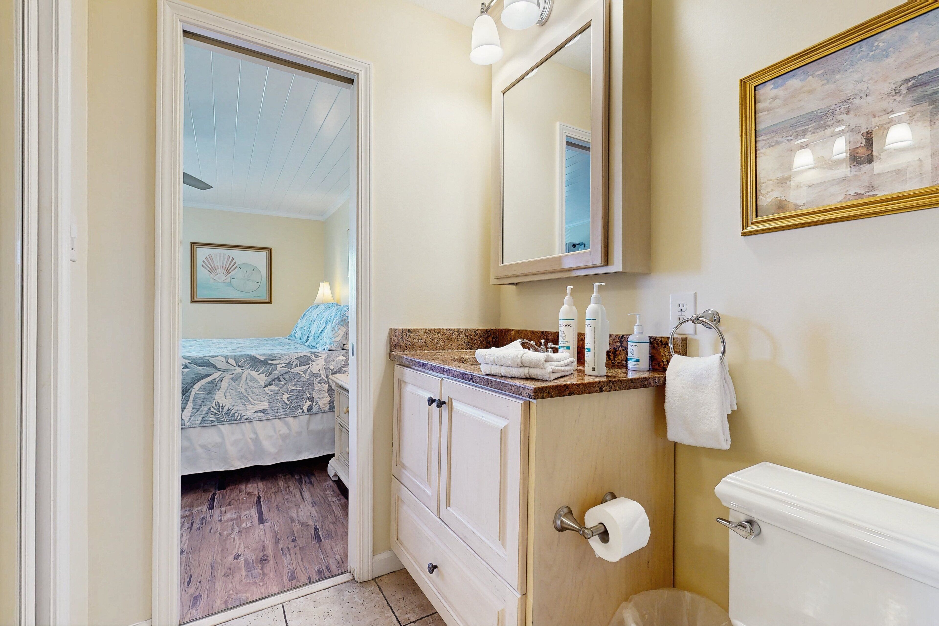 A bathroom with a toilet, sink, and mirror. There is a bed visible through an open door in the adjacent room.