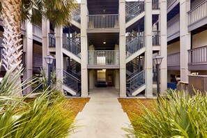 A multi-story building with a central atrium, balconies, and a pathway flanked by plants. - Ocean-view Condo With Pool, Balcony, AC & W/D - Walk to Beach & Near Downtown (Fernandina Beach)