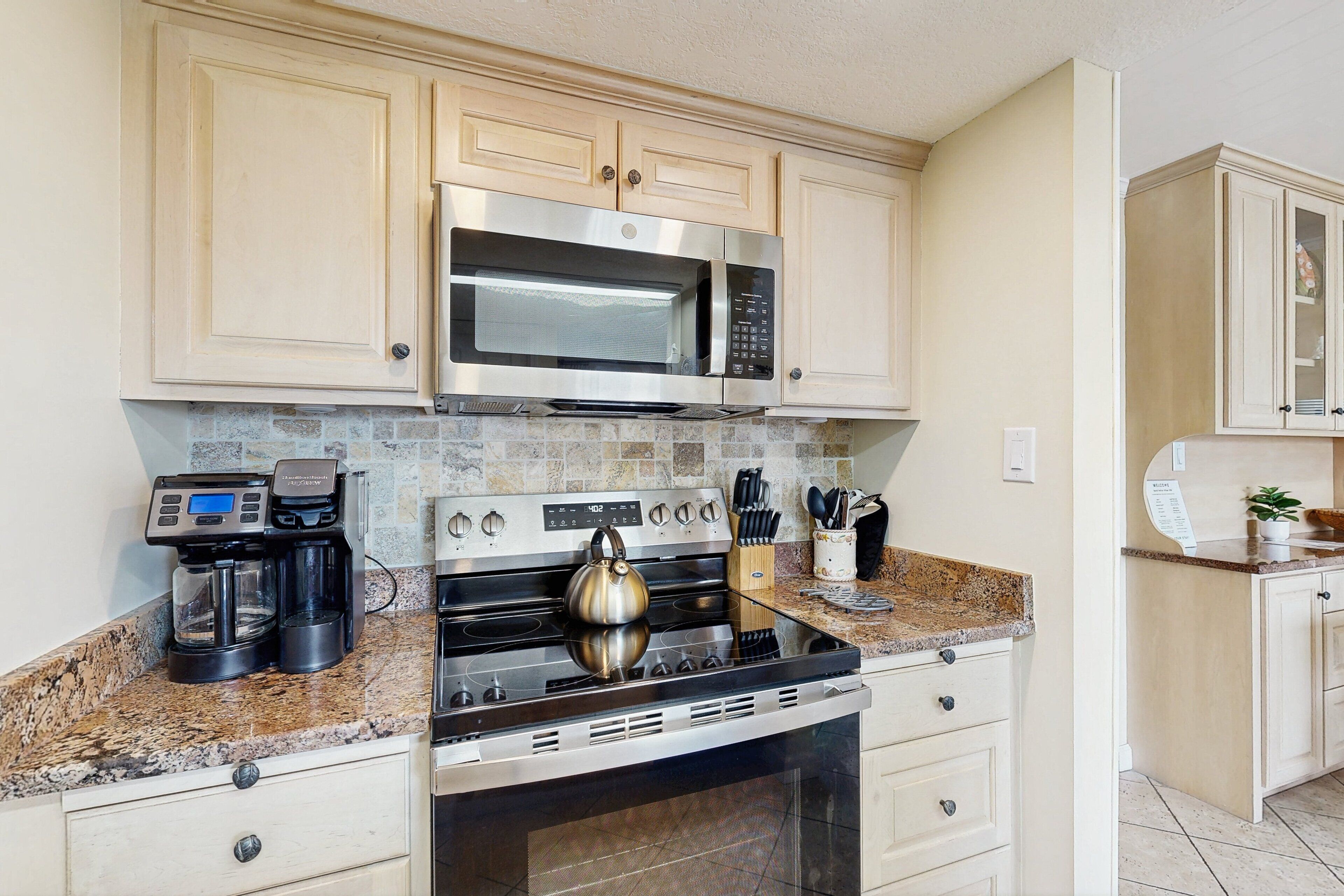 A kitchen with light-colored cabinets, a stainless steel microwave, a coffee maker, and a kettle on the countertop.