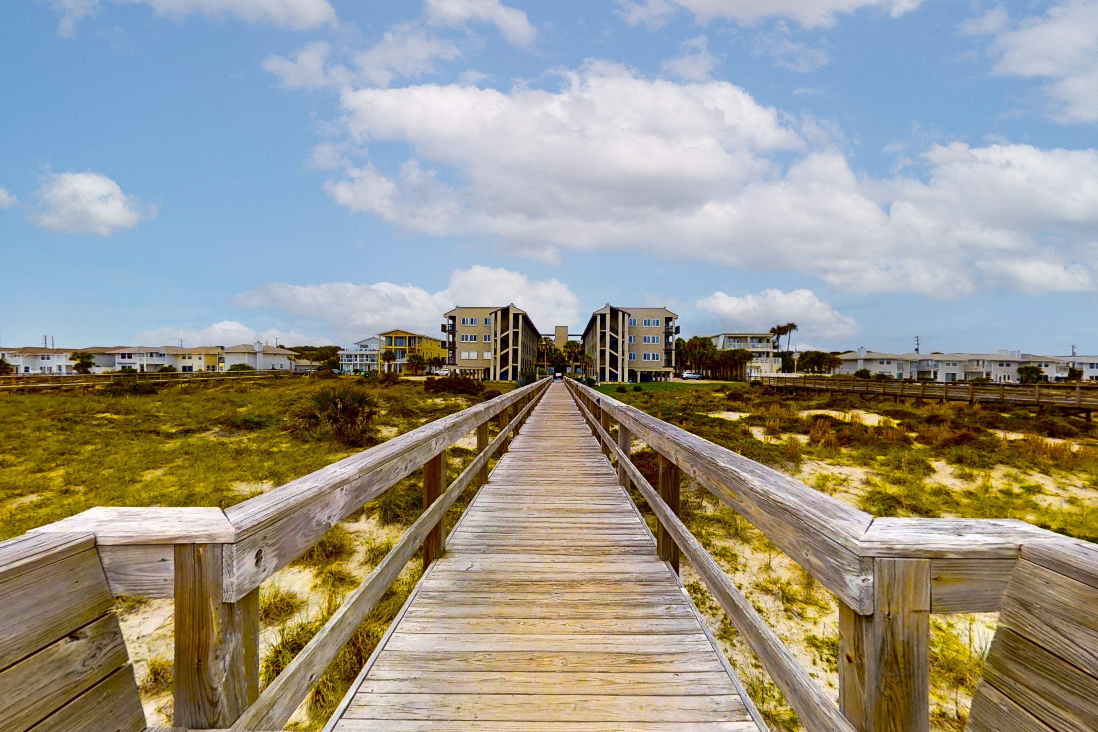 A wooden boardwalk leading to a beachfront area with buildings in the background.