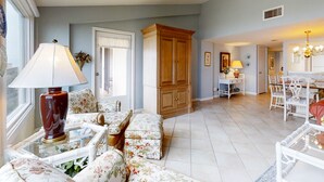 A living room with floral-patterned sofas, a wooden cabinet, and a dining area with white chairs and a chandelier. - Oceanfront Condo With Spacious Balcony, Great Views, AC & Outdoor Pooloc (Fernandina Beach)
