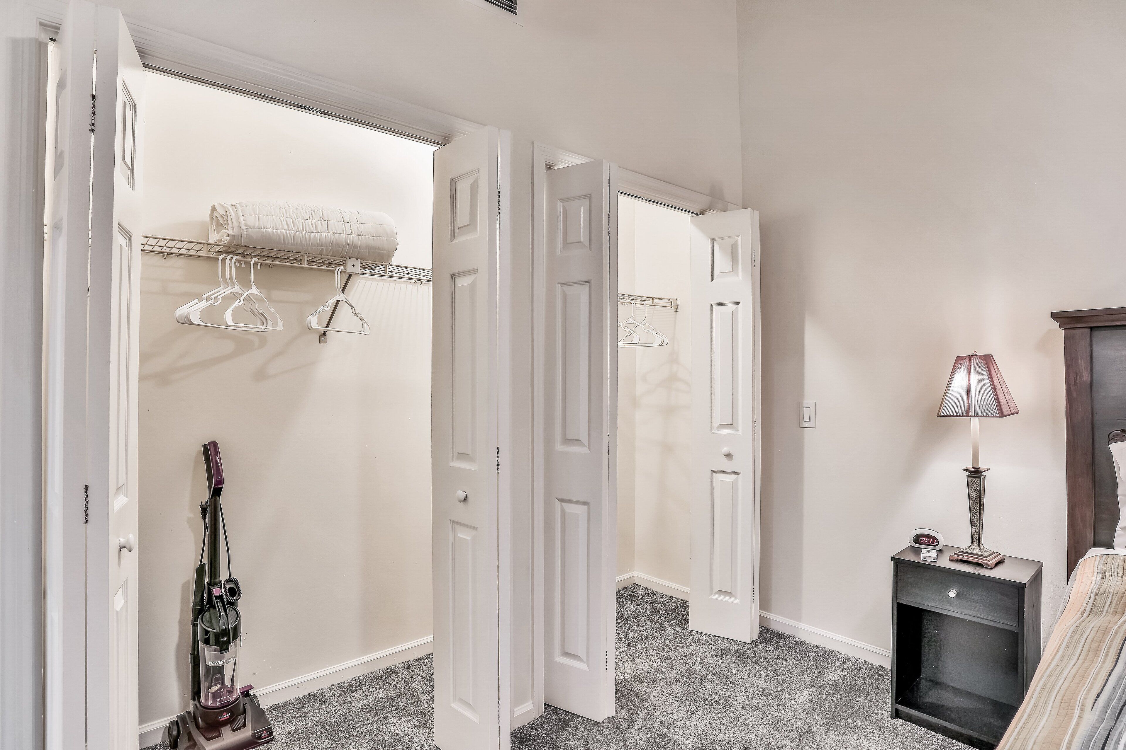 A hallway with a vacuum cleaner, open doors, a coat rack, a lamp, and a bed.