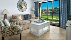 Un salon avec un canapé beige, un fauteuil à motifs et une table basse blanche. Un grand miroir rond est accroché au mur, et une grande fenêtre offre une vue sur un paysage verdoyant.