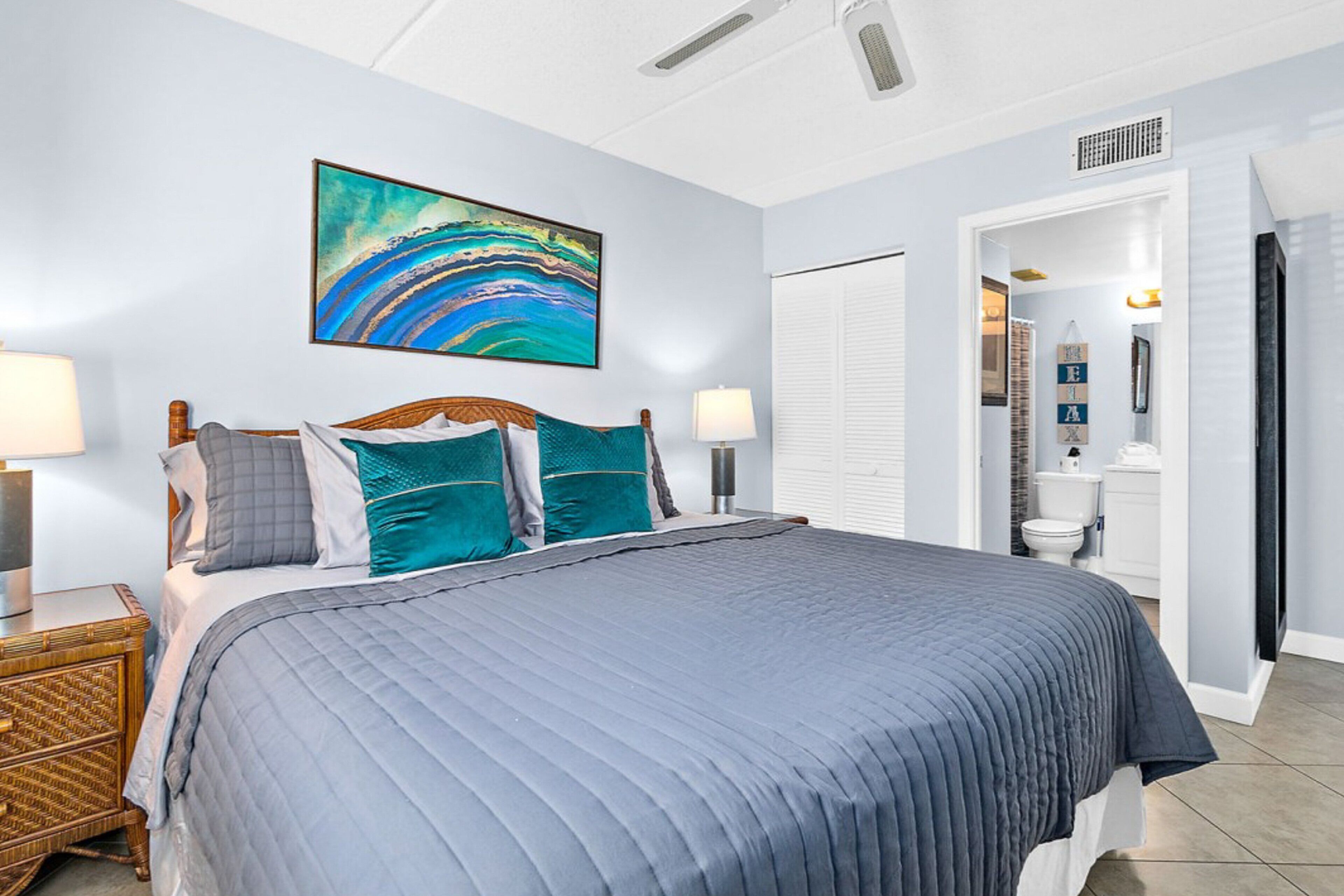 Ein Schlafzimmer mit einem großen Bett, zwei Nachttischen, einem Wandbild und einem durch eine offene Tür einsehbaren Badezimmer.