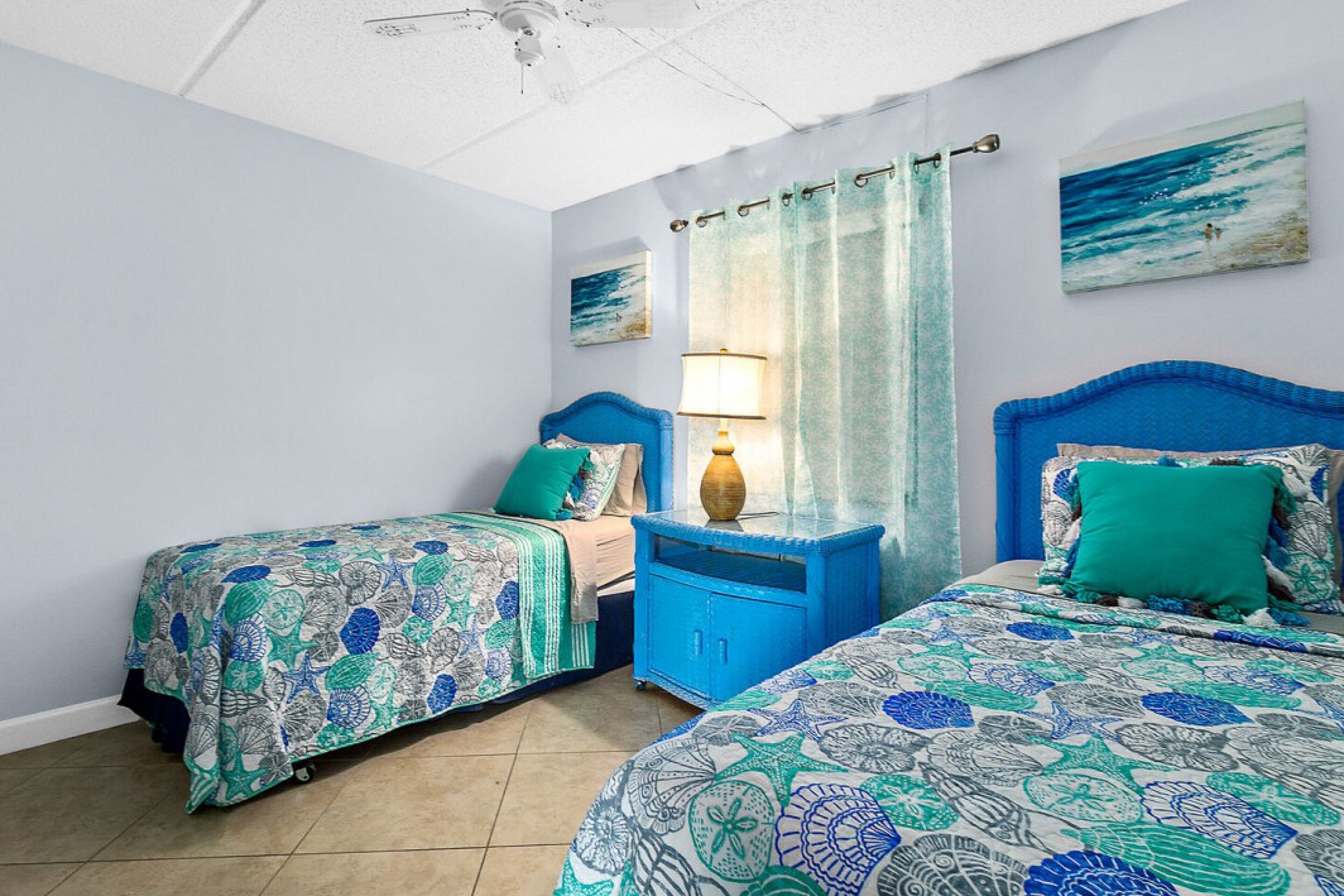 A bedroom with two beds, each with a blue headboard, a nightstand with a lamp, and a window with a curtain.