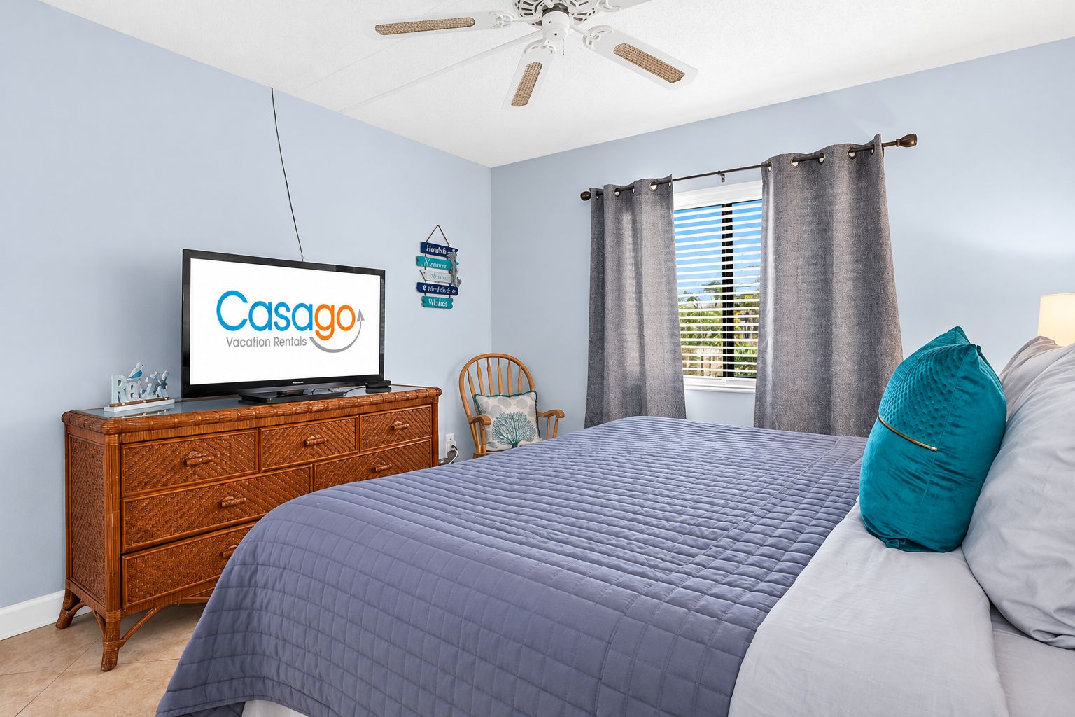 Un dormitorio con una cama, televisión, cómoda de madera, una silla y una ventana con cortinas.