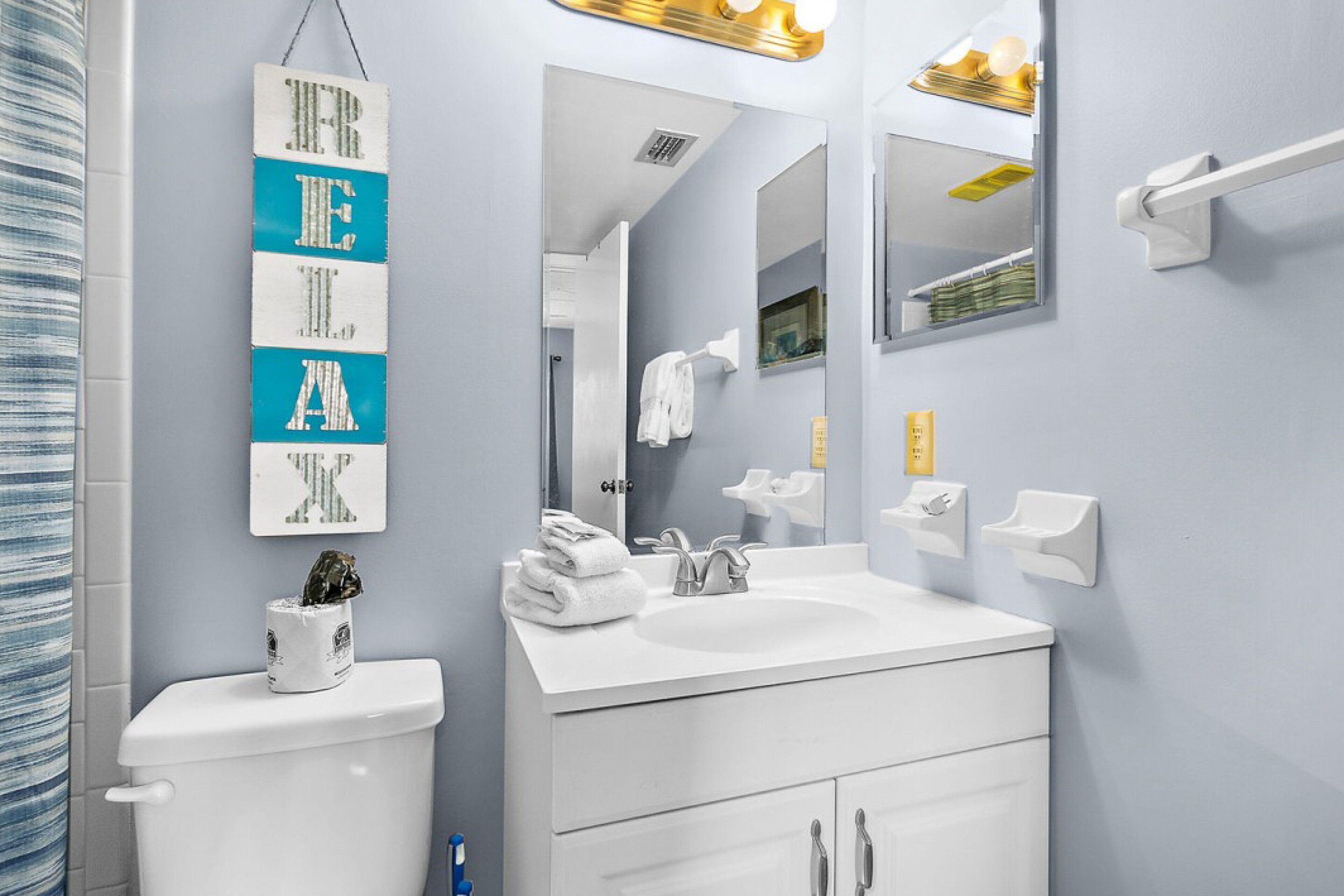 Une salle de bain moderne avec un lavabo blanc, un miroir et des toilettes. On y trouve un porte-serviettes avec des serviettes pliées et une petite plante en pot posée sur les toilettes.