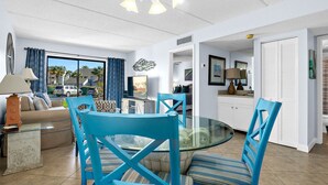 Un salon moderne avec une table à manger en verre, des chaises bleues, un canapé et un coin cuisine visible à travers une porte ouverte.